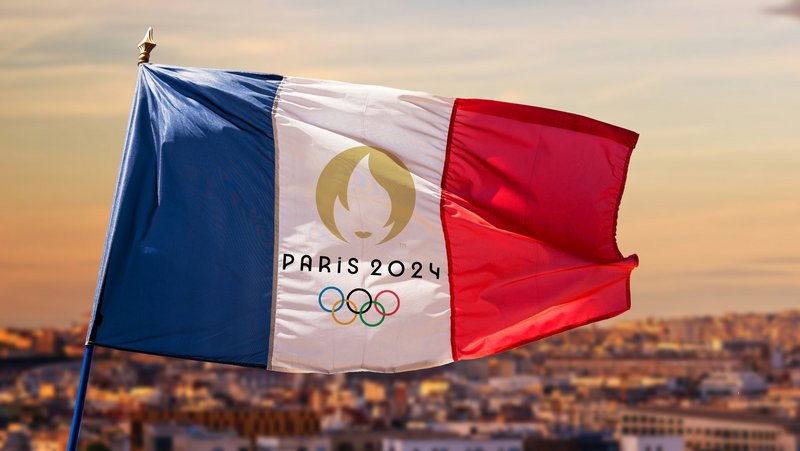 Das Foto zeigt die französiche Flagge mit Olympia 2024 Aufdruck vor der Parioser Skyline mit dem Eifeltum im Hintergrund